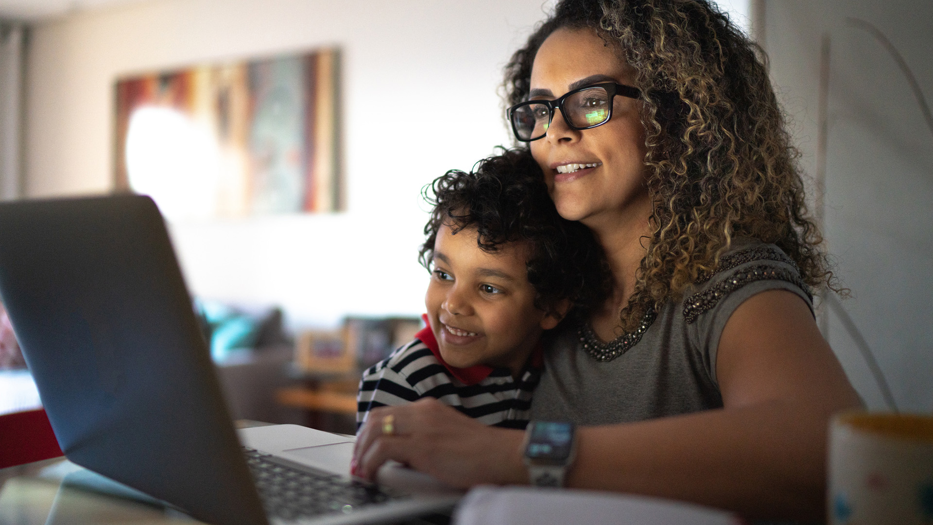 A imagem mostra uma mulher, com uma criança no colo, na frente de um notebook. São pessoas pretas e demonstram alegria. A imagem quer representar como a mulher lida com a maternidade e carreira.