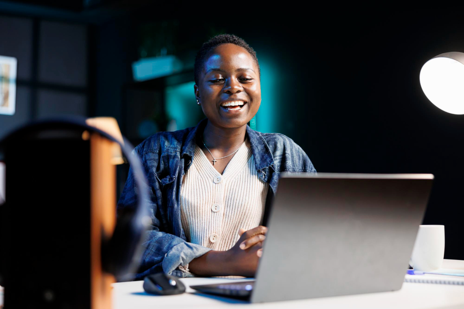 A imagem mostra uma mulher negra retinta, usando uma camisa branca e uma jaqueta jeans, sorrindo, em frente a um notebook. A imagem representa uma analista de marketing sênior.