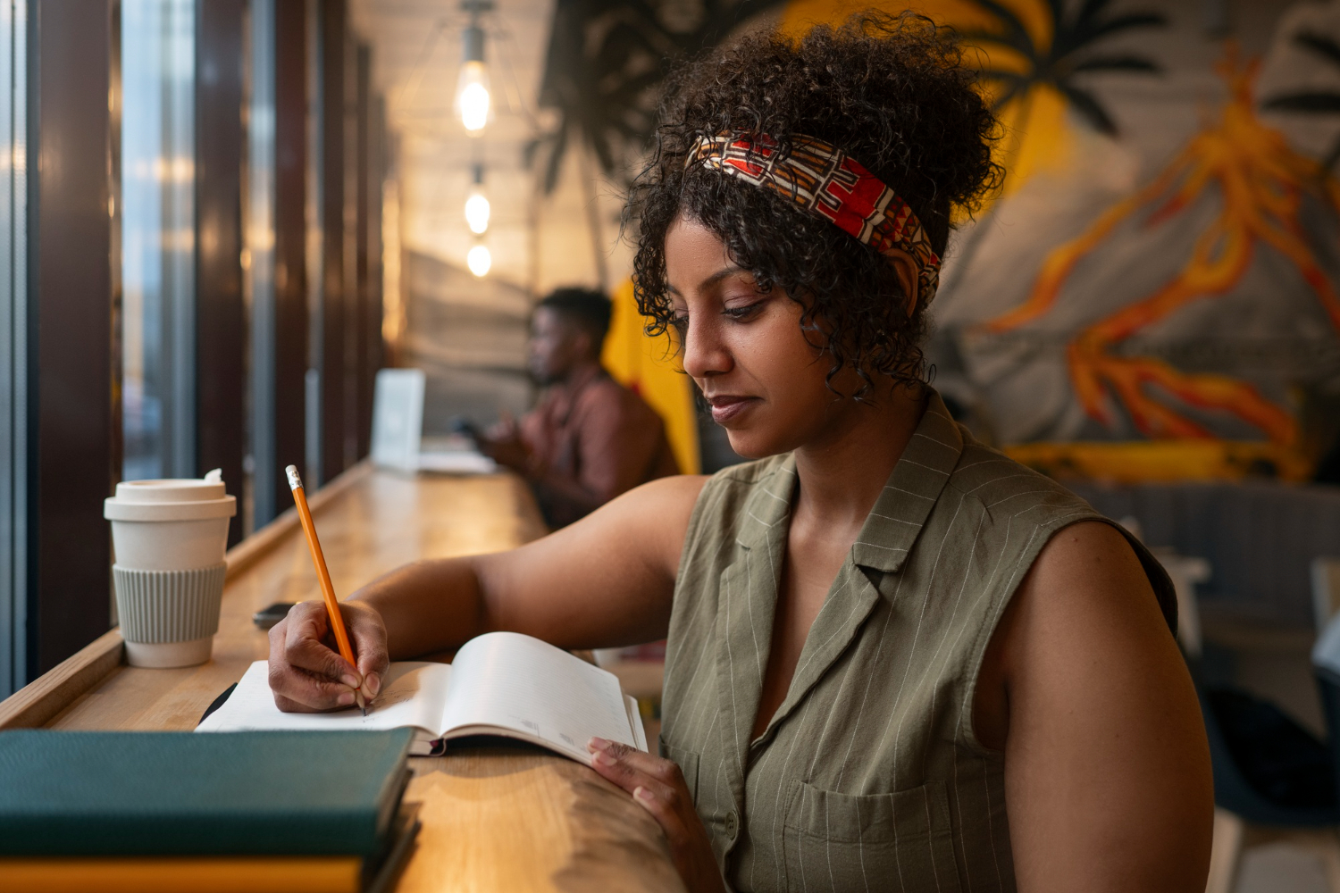 Mulher preta, com cabelo cacheado, usando uma faixa estampada, sentada em um ambiente de cafeteria, escrevendo em seu caderno com foco. Ao lado dela, há uma xícara de café reutilizável. A imagem reflete um momento de planejamento semanal, em que ela organiza suas atividades com concentração e determinação, aproveitando o ambiente acolhedor para se preparar para os desafios da semana