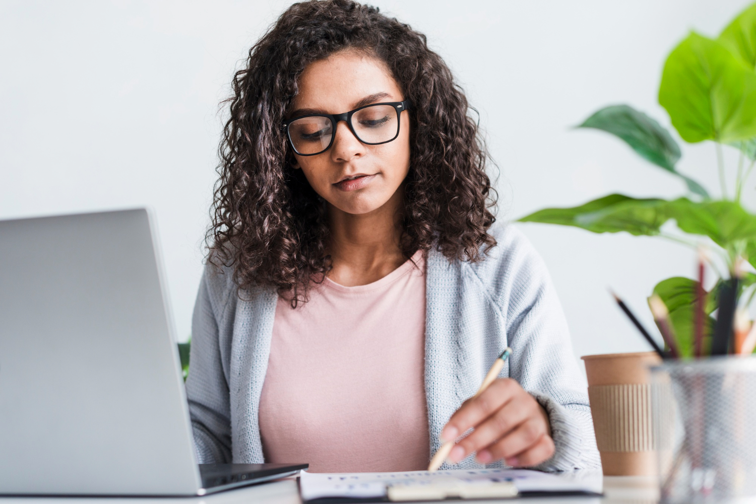 Redatora publicitária concentrada em seu trabalho, revisando anotações enquanto utiliza um laptop. A profissional, usando óculos e com cabelos cacheados, está sentada em uma mesa de trabalho, em um ambiente claro e organizado, refletindo foco e criatividade