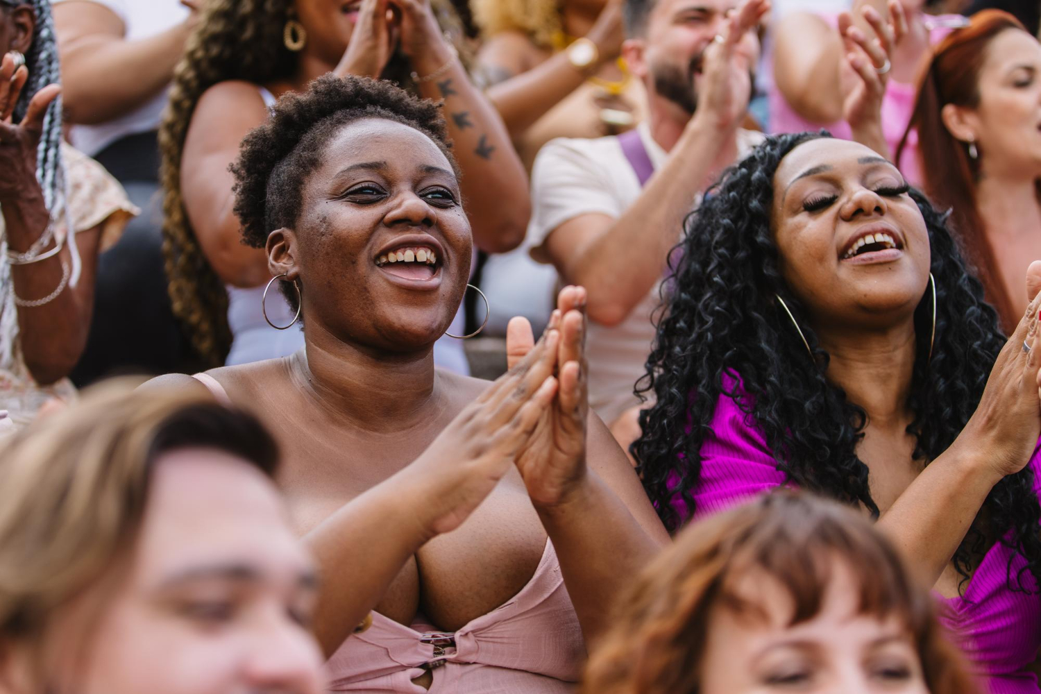 Uma multidão diversa participa de um evento ao ar livre, onde duas mulheres negras, sorridentes e demonstrando entusiasmo, aplaudem com energia. O ambiente é alegre e acolhedor, refletindo o acesso e a inclusão de mulheres negras e indígenas em espaços de destaque e celebração. A imagem ressalta o empoderamento feminino e a diversidade, com foco na representatividade de mulheres de diferentes origens étnicas em eventos públicos e culturais