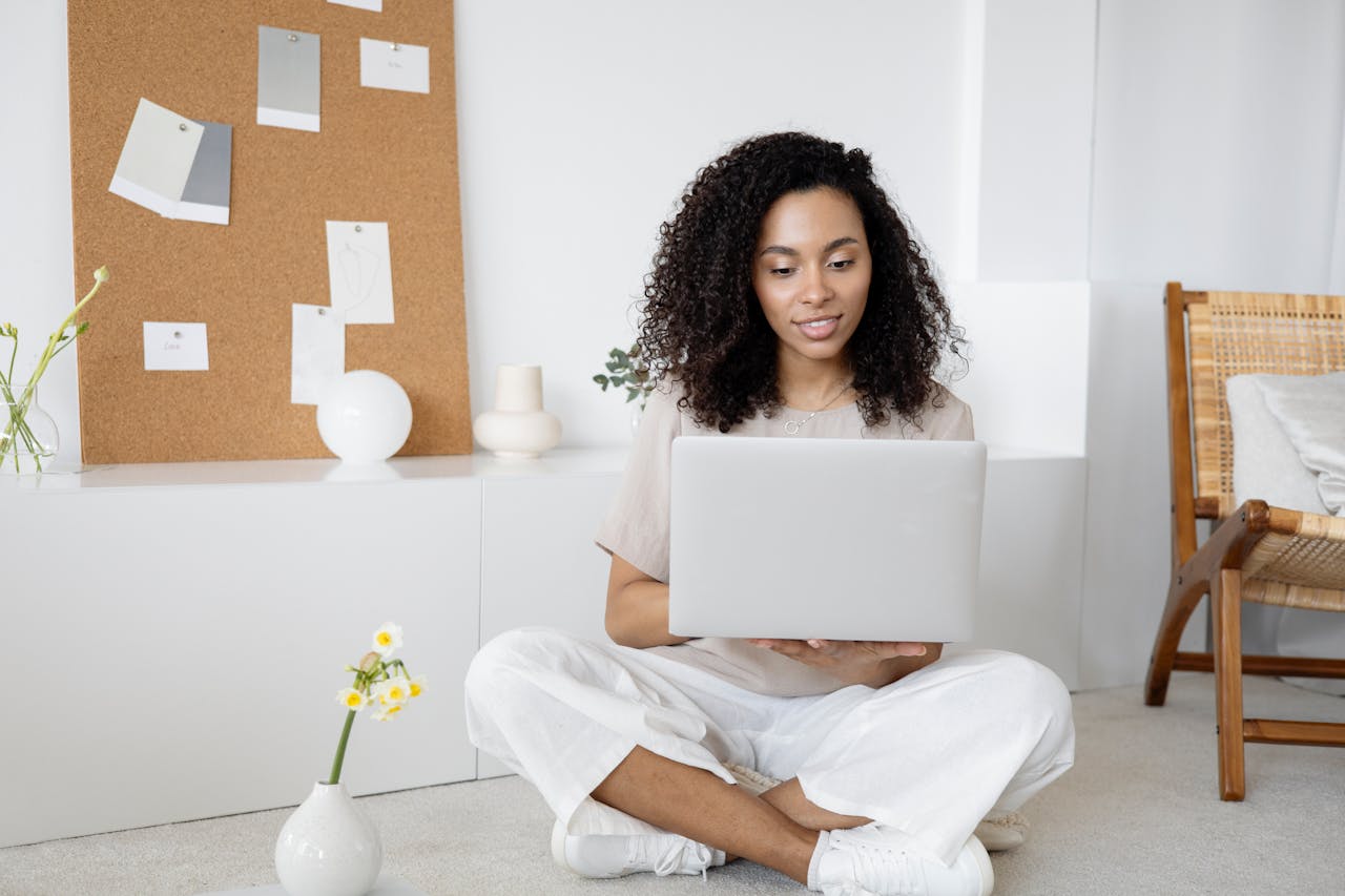 Cena de um ambiente minimalista, onde uma mulher está sentada no chão usando um notebook, representando um momento de planejamento e criação. Ao fundo, um painel de cortiça com recortes e amostras sugere inspiração visual e organização de ideias. O espaço harmonioso, com tons neutros e elementos decorativos simples, reflete um ambiente propício para o desenvolvimento de estratégias de branding criativas e focadas.