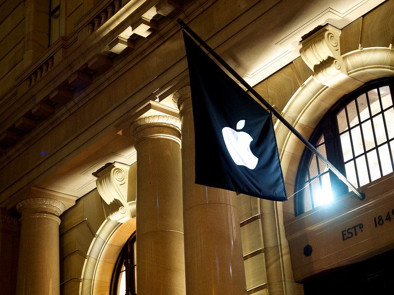 Fachada de um edifício histórico com colunas imponentes, iluminada por luzes amarelas à noite. Em destaque, uma bandeira preta com o logotipo branco da Apple está hasteada, contrastando com a arquitetura clássica do local. Na lateral, a inscrição 'EST. 1841' pode ser vista em relevo na parede.