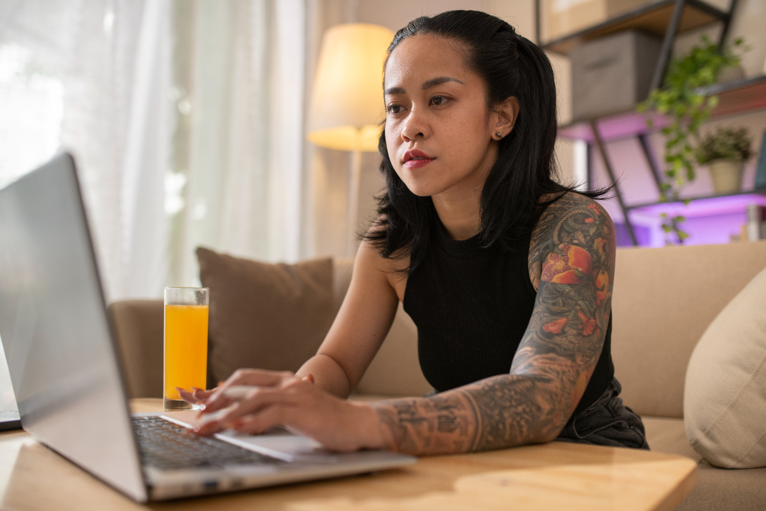 Uma mulher de origem indígena, concentrada trabalha em um laptop em um ambiente acolhedor, com uma luminária acesa ao fundo e um copo de suco na mesa. A cena reflete a criatividade e o foco necessários no desenvolvimento de estratégias de branding digital, destacando o uso de ferramentas online para fortalecer a identidade de marcas no ambiente virtual.