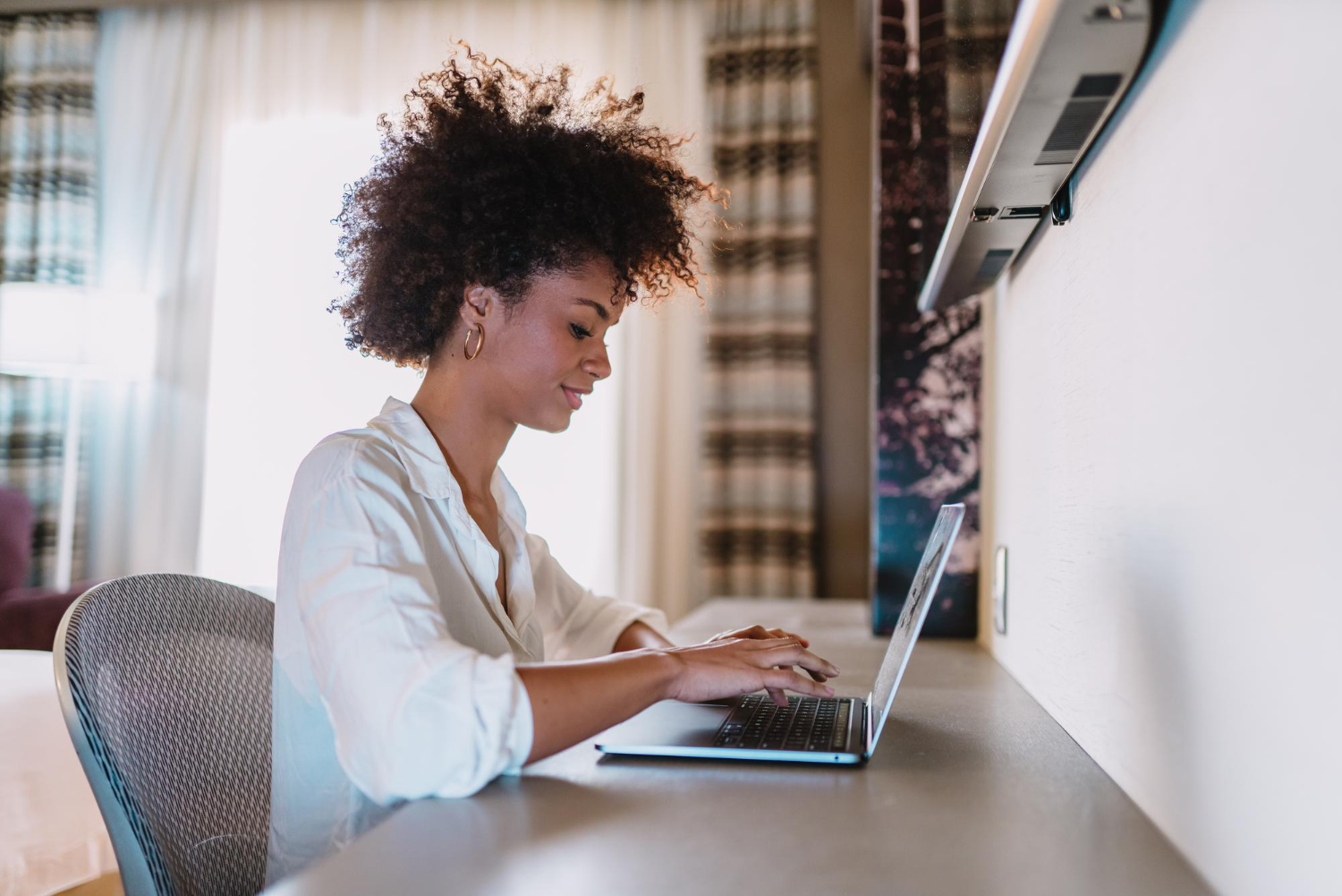 Uma mulher jovem, com cabelo crespo volumoso, trabalha concentrada em um notebook em um ambiente moderno e iluminado. Vestindo uma camisa branca, ela sorri levemente enquanto digita, transmitindo confiança e tranquilidade. O cenário inclui uma cadeira ergonômica e cortinas elegantes ao fundo, sugerindo um espaço confortável e produtivo. A imagem simboliza o empoderamento digital, destacando a autonomia e a conexão da mulher com a tecnologia para trabalhar ou se desenvolver profissionalmente em um ambiente virtual.