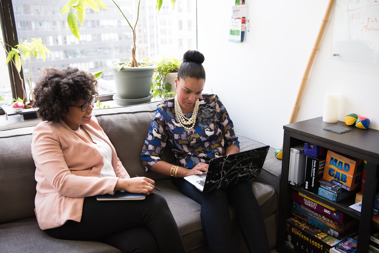 Duas mulheres sentadas em um sofá de escritório discutem ideias enquanto olham para um laptop. Uma delas usa um blazer rosa e segura um caderno, enquanto a outra usa uma blusa estampada e um colar de pérolas. Ao fundo, há plantas, uma prateleira com jogos e livros, e uma lousa branca com anotações. A cena reflete um ambiente colaborativo e acolhedor, simbolizando como marcas com propósito podem criar espaços que incentivam criatividade, inclusão e impacto positivo.