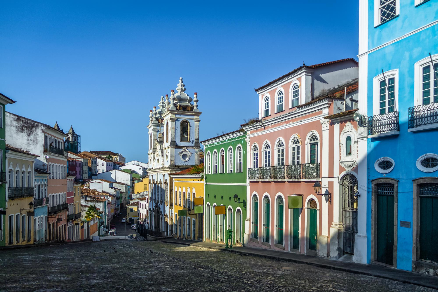 Rua do Pelourinho, em Salvador, Bahia, com seu calçamento de pedra e casarões coloridos que refletem a riqueza da arquitetura colonial brasileira. Ao fundo, a igreja barroca de fachada branca se destaca, representando a mistura de culturas e a ancestralidade presente nas influências africanas, europeias e indígenas que moldaram a história e o patrimônio cultural da região. O céu azul complementa a atmosfera vibrante e histórica do local.