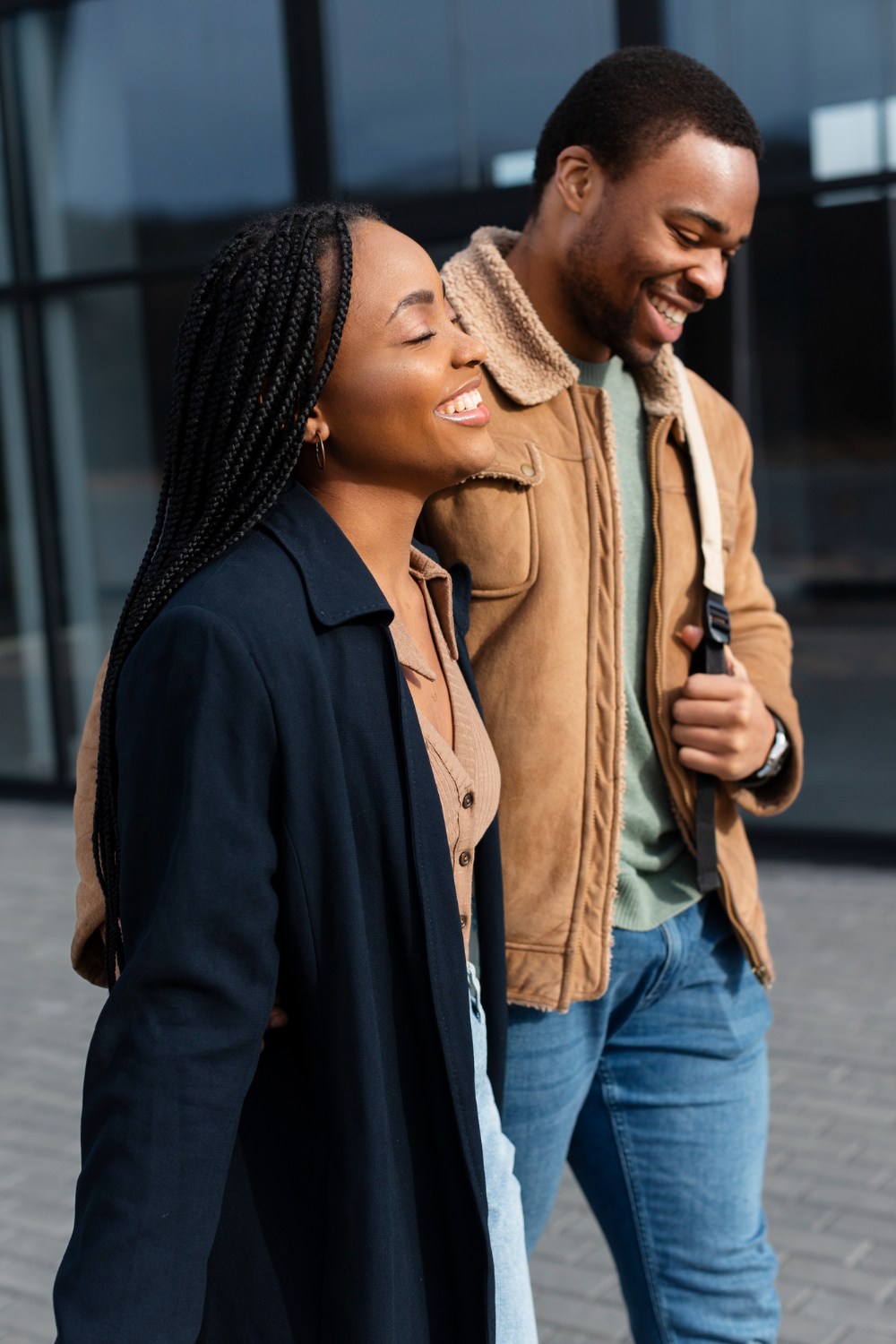 Duas personas reais interagem de forma descontraída enquanto caminham ao ar livre. Uma mulher, com tranças longas e sorriso radiante, veste um blazer azul escuro e uma blusa bege. Ao seu lado, um homem também sorri enquanto usa uma jaqueta de camurça com detalhes de lã, jeans e uma bolsa a tiracolo. Ambos transmitem alegria e espontaneidade, representando momentos autênticos e naturais de convivência. Ao fundo, há um prédio de vidro que complementa o cenário urbano da cena.