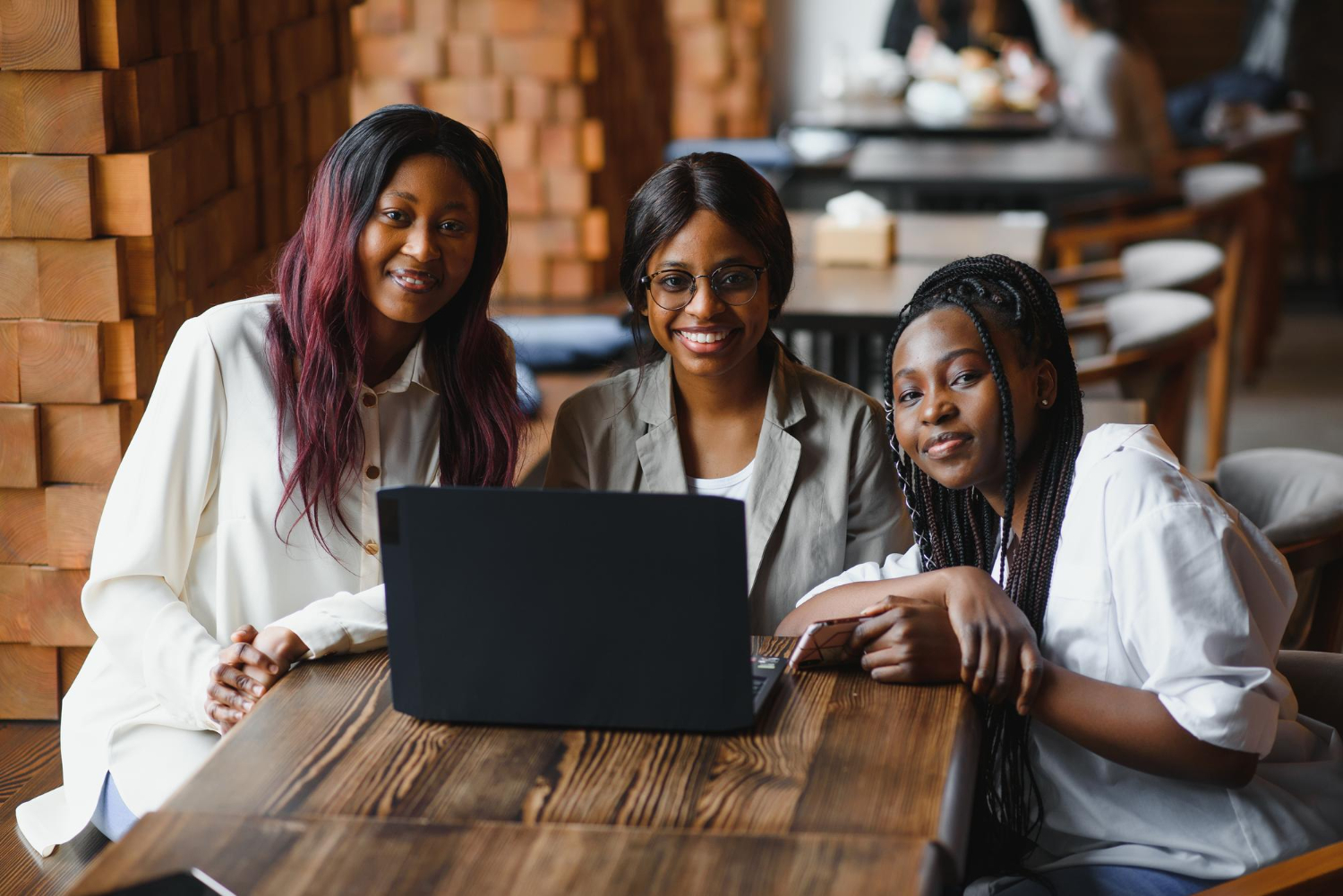 Três mulheres sentadas ao redor de uma mesa de madeira, sorrindo e olhando para a câmera, com um laptop aberto à frente delas. O ambiente é moderno e acolhedor, com mesas e cadeiras ao fundo. A imagem transmite colaboração e trabalho em equipe, representando o conceito de planejamento de marketing digital, destacando criatividade, estratégia e parceria no desenvolvimento de soluções para alcançar resultados online.