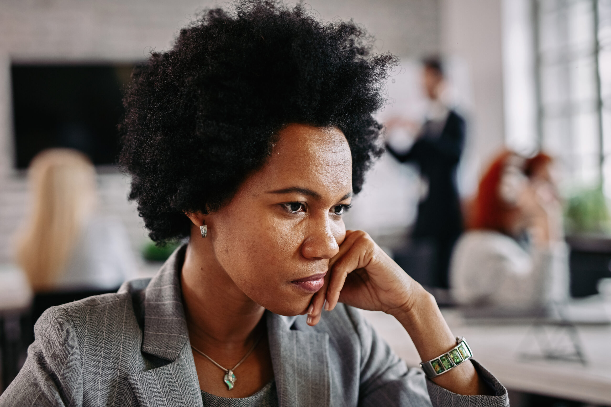 Mulher de expressão pensativa está em um ambiente de trabalho, apoiando o rosto na mão enquanto reflete. Ela veste um blazer cinza e acessórios discretos, demonstrando seriedade e introspecção. Ao fundo, outras pessoas aparecem desfocadas, simbolizando a sensação de isolamento em meio a um contexto social. A imagem ilustra o conceito de "solidão na carreira," refletindo os desafios emocionais e as responsabilidades individuais de posições de liderança.