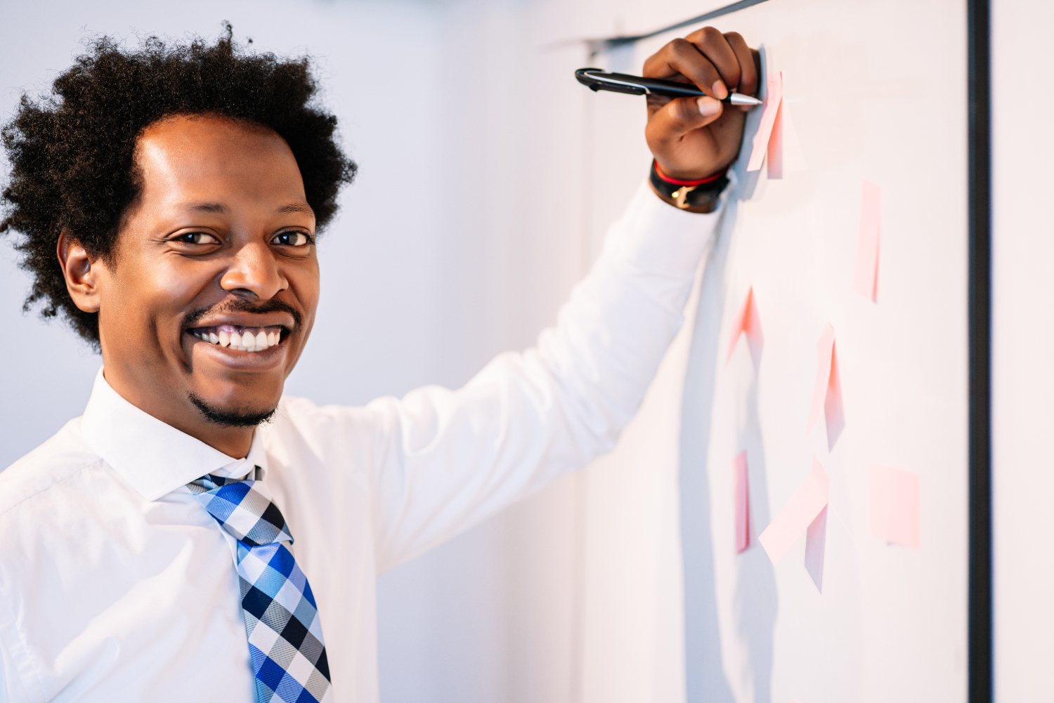 Homem preto, sorridente, de camisa social branca e gravata xadrez azul, está escrevendo ou fixando notas adesivas em um quadro branco. Ele segura uma caneta enquanto olha para a câmera, transmitindo confiança e entusiasmo. As notas adesivas no quadro sugerem organização e brainstorming. A imagem remete ao conceito de storytelling, destacando a construção de narrativas visuais e verbais de forma criativa para engajar e inspirar audiências.