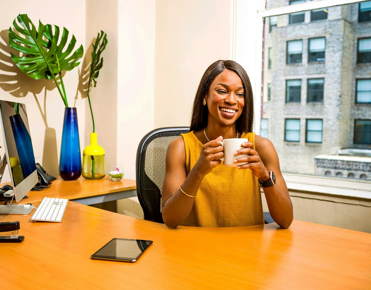 Mulher sorridente sentada em uma mesa de escritório bem iluminada, segurando uma xícara de café. No ambiente, há elementos de decoração modernos, como vasos coloridos com plantas, um computador e um tablet sobre a mesa. Ao fundo, uma grande janela exibe prédios urbanos, transmitindo um ar profissional e acolhedor. A imagem ilustra o conceito de valor percebido, destacando conforto, bem-estar e produtividade em um ambiente de trabalho inspirador.