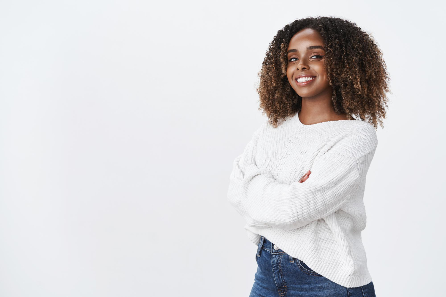 Uma mulher preta sorridente com cabelos cacheados volumosos e mechas douradas, vestindo um suéter branco e calça jeans, posa com os braços cruzados em um fundo branco. Sua expressão confiante reflete autoestima e segurança, transmitindo uma mensagem de força e positividade.