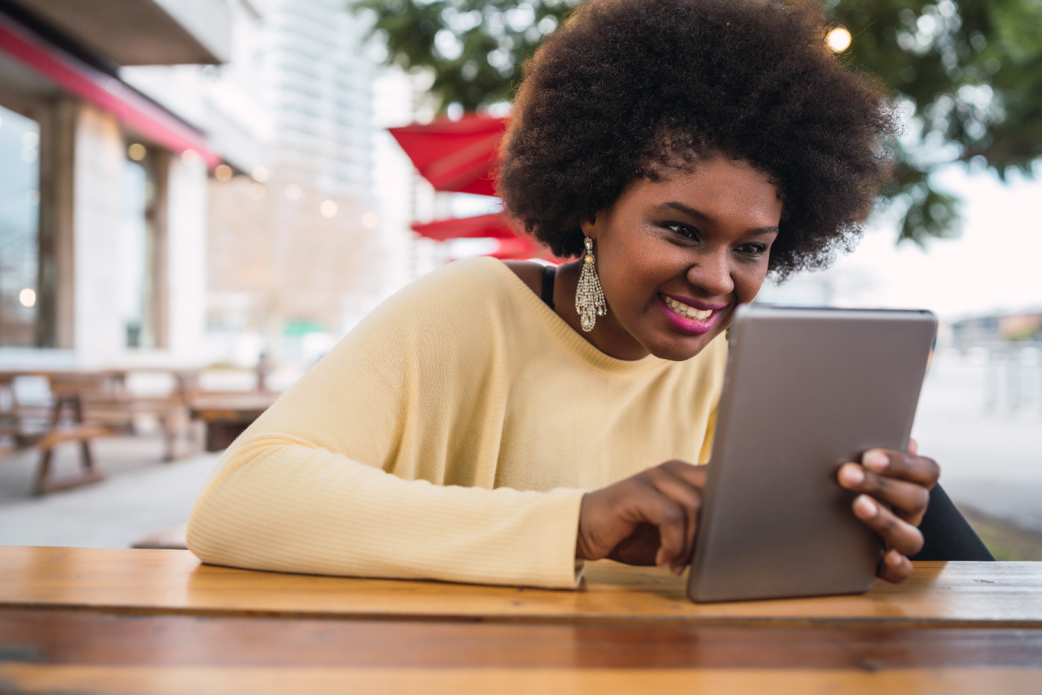 Mulher sorridente com cabelo afro e blusa amarela, sentada em uma mesa ao ar livre em um ambiente urbano. Ela segura um tablet com as duas mãos e interage com a tela de forma esportiva e liberada, demonstrando um comportamento relaxado e positivo.