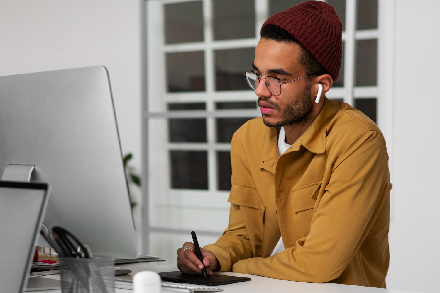 Homem jovem preto com barba fina, usando óculos, fones de ouvido brancos e um gorro bordô, trabalha concentrado em frente a um computador. Ele segura uma caneta digital enquanto desenha em uma mesa gráfica, em um ambiente de escritório moderno. A cena sugere um profissional de design trabalhando na criação de uma identidade visual.