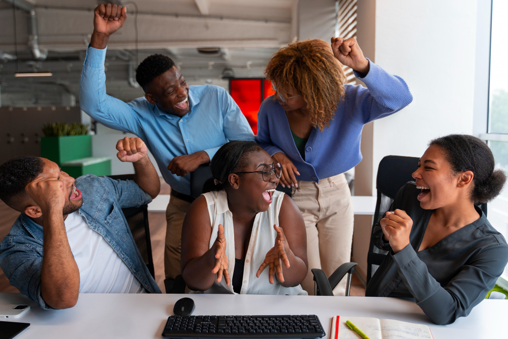 dois homens negros e 3 mulheres negras estão em um ambiente corporativo e comemoram algo em torno de uma mesa de escritório