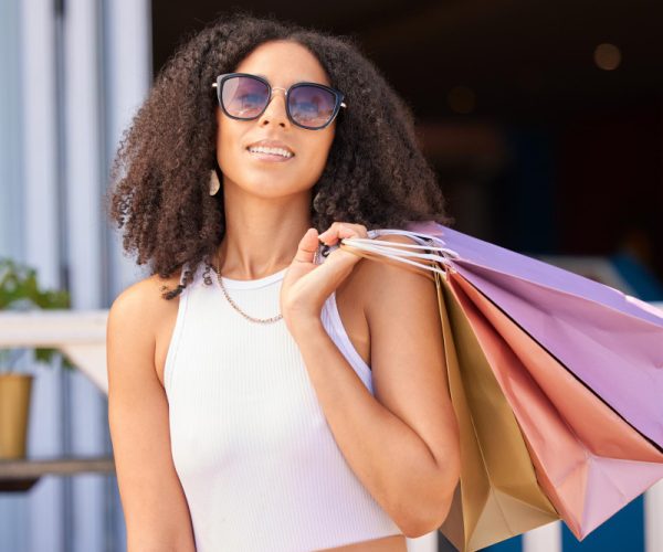 mulher negra com cabelo médio bem cacheado., vestindo camiseta branca e óculos escuro, segura com a mão direta por trás dos ombros três sacolas de compras coloridas