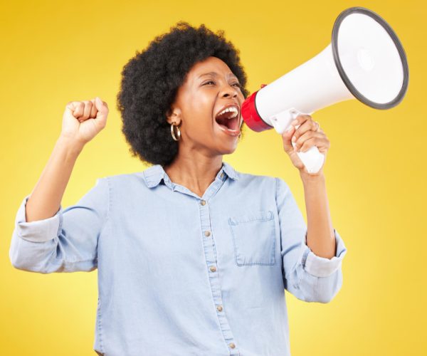 mulher negra vestindo camiseta jeans, faz símbolo de resistência com a mão esquerda e com a mão direita segura um megafone.