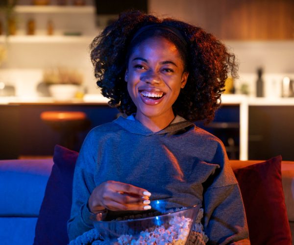 jovem negra usando moletom, com o black power solto, sorri sentada em um sofá vendo streaming e come pipoca.
