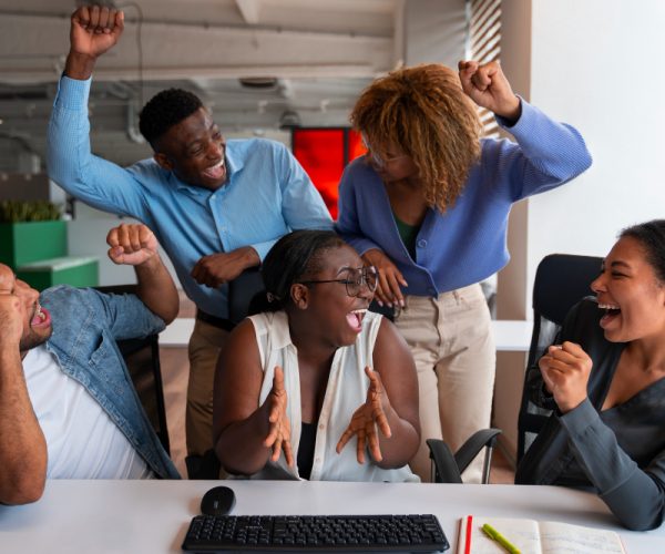 dois homens negros e 3 mulheres negras estão em um ambiente corporativo e comemoram algo em torno de uma mesa de escritório