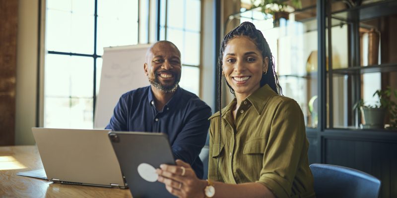 Female executive and bearded colleague working with digital devices, smiling cheerfully at camera in boardroom