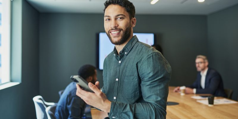 A confident businessman wearing a casual shirt leans on a conference table while holding his phone, with colleagues discussing work in the background.