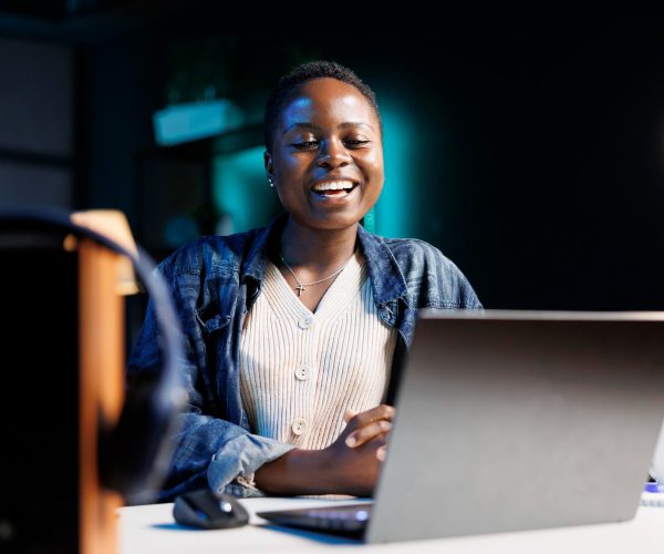 A imagem mostra uma mulher negra retinta, usando uma camisa branca e uma jaqueta jeans, sorrindo, em frente a um notebook. A imagem representa uma analista de marketing sênior.