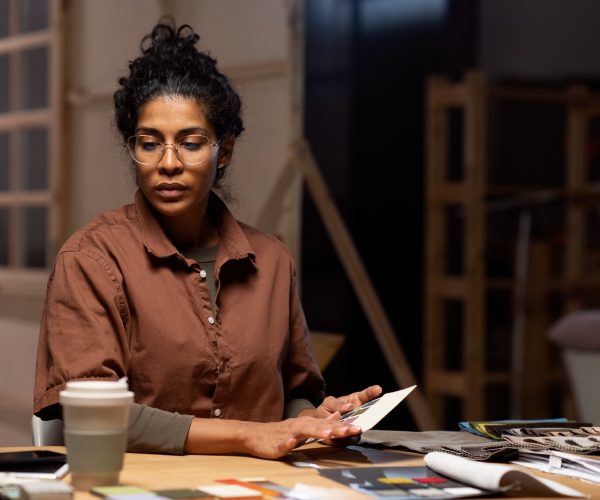 Mulher trabalhando em um ambiente criativo, rodeada por amostras de tecidos, papéis e materiais de design. Ela está concentrada, segurando uma paleta de cores, enquanto parece planejar algo relacionado à identidade visual. O espaço ao fundo tem um estilo rústico, reforçando o conceito de criação artesanal e detalhada, ideal para um trabalho como 'Arquitetura de Marca'.