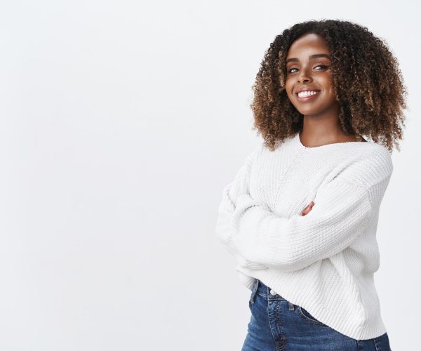 Uma mulher preta sorridente com cabelos cacheados volumosos e mechas douradas, vestindo um suéter branco e calça jeans, posa com os braços cruzados em um fundo branco. Sua expressão confiante reflete autoestima e segurança, transmitindo uma mensagem de força e positividade.