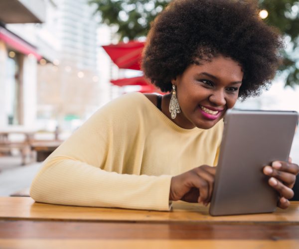 Mulher sorridente com cabelo afro e blusa amarela, sentada em uma mesa ao ar livre em um ambiente urbano. Ela segura um tablet com as duas mãos e interage com a tela de forma esportiva e liberada, demonstrando um comportamento relaxado e positivo.