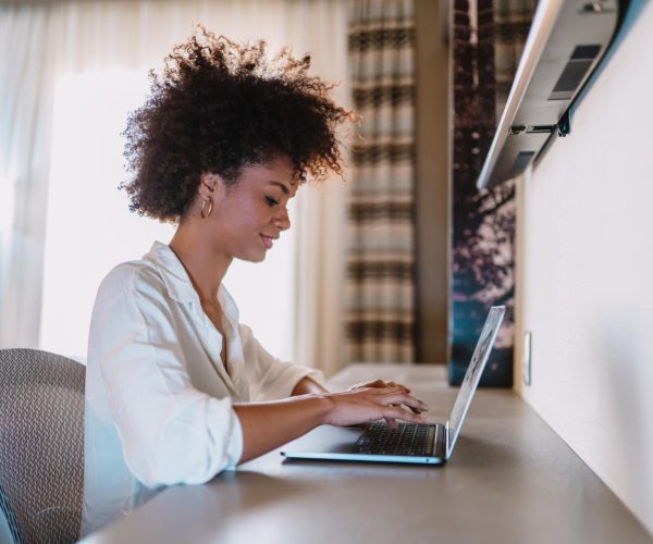 Uma mulher jovem, com cabelo crespo volumoso, trabalha concentrada em um notebook em um ambiente moderno e iluminado. Vestindo uma camisa branca, ela sorri levemente enquanto digita, transmitindo confiança e tranquilidade. O cenário inclui uma cadeira ergonômica e cortinas elegantes ao fundo, sugerindo um espaço confortável e produtivo. A imagem simboliza o empoderamento digital, destacando a autonomia e a conexão da mulher com a tecnologia para trabalhar ou se desenvolver profissionalmente em um ambiente virtual.