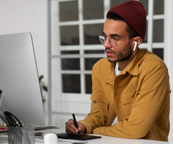 Homem jovem preto com barba fina, usando óculos, fones de ouvido brancos e um gorro bordô, trabalha concentrado em frente a um computador. Ele segura uma caneta digital enquanto desenha em uma mesa gráfica, em um ambiente de escritório moderno. A cena sugere um profissional de design trabalhando na criação de uma identidade visual.