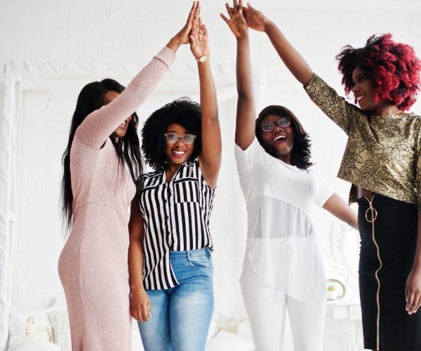 Quatro mulheres celebrando juntas em um ambiente claro e acolhedor. Elas estão com os braços levantados, unindo as mãos no alto, simbolizando união e conquistas. A diversidade de estilos e expressões transmite energia positiva e determinação, representando um momento de celebração coletiva. Essa imagem pode ilustrar o conceito de uma jornada 2024 repleta de desafios superados e metas alcançadas, com foco na colaboração e no empoderamento.