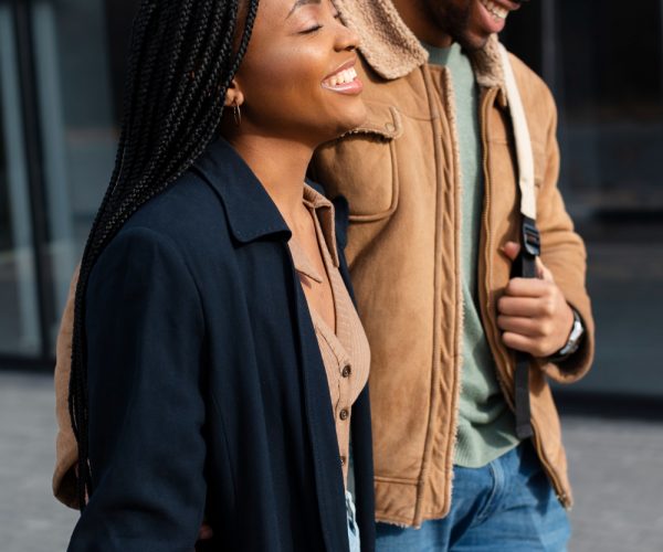 Duas personas reais interagem de forma descontraída enquanto caminham ao ar livre. Uma mulher, com tranças longas e sorriso radiante, veste um blazer azul escuro e uma blusa bege. Ao seu lado, um homem também sorri enquanto usa uma jaqueta de camurça com detalhes de lã, jeans e uma bolsa a tiracolo. Ambos transmitem alegria e espontaneidade, representando momentos autênticos e naturais de convivência. Ao fundo, há um prédio de vidro que complementa o cenário urbano da cena.