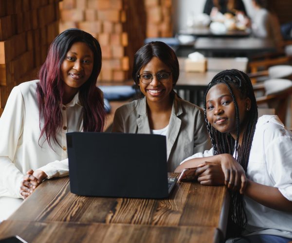 Três mulheres sentadas ao redor de uma mesa de madeira, sorrindo e olhando para a câmera, com um laptop aberto à frente delas. O ambiente é moderno e acolhedor, com mesas e cadeiras ao fundo. A imagem transmite colaboração e trabalho em equipe, representando o conceito de planejamento de marketing digital, destacando criatividade, estratégia e parceria no desenvolvimento de soluções para alcançar resultados online.