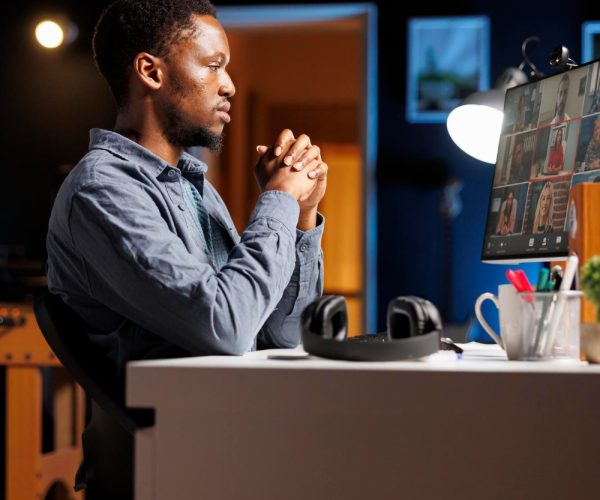 Um homem preto está sentado à mesa em um ambiente de trabalho, participando de uma reunião virtual exibida em seu monitor. Ele está com as mãos entrelaçadas, demonstrando atenção e reflexão. Ao lado, fones de ouvido e materiais de escritório reforçam o foco em estratégias colaborativas. A cena representa o planejamento estratégico envolvido no processo de rebranding, evidenciando a importância da comunicação visual e da redefinição de identidade para fortalecer marcas.