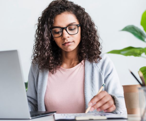 Redatora publicitária concentrada em seu trabalho, revisando anotações enquanto utiliza um laptop. A profissional, usando óculos e com cabelos cacheados, está sentada em uma mesa de trabalho, em um ambiente claro e organizado, refletindo foco e criatividade