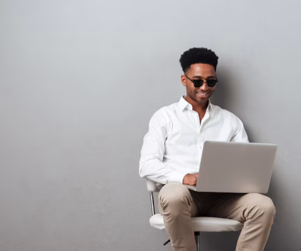 homem negro sentado vestido camisa branca e calça pege trabalha com um computador descansando nas pernas.