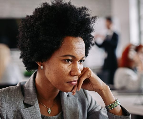 Mulher de expressão pensativa está em um ambiente de trabalho, apoiando o rosto na mão enquanto reflete. Ela veste um blazer cinza e acessórios discretos, demonstrando seriedade e introspecção. Ao fundo, outras pessoas aparecem desfocadas, simbolizando a sensação de isolamento em meio a um contexto social. A imagem ilustra o conceito de "solidão na carreira," refletindo os desafios emocionais e as responsabilidades individuais de posições de liderança.