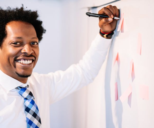 Homem preto, sorridente, de camisa social branca e gravata xadrez azul, está escrevendo ou fixando notas adesivas em um quadro branco. Ele segura uma caneta enquanto olha para a câmera, transmitindo confiança e entusiasmo. As notas adesivas no quadro sugerem organização e brainstorming. A imagem remete ao conceito de storytelling, destacando a construção de narrativas visuais e verbais de forma criativa para engajar e inspirar audiências.