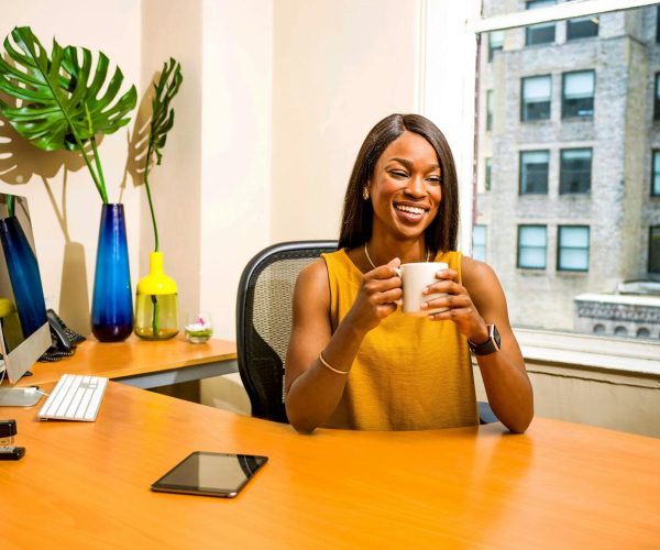 Mulher sorridente sentada em uma mesa de escritório bem iluminada, segurando uma xícara de café. No ambiente, há elementos de decoração modernos, como vasos coloridos com plantas, um computador e um tablet sobre a mesa. Ao fundo, uma grande janela exibe prédios urbanos, transmitindo um ar profissional e acolhedor. A imagem ilustra o conceito de valor percebido, destacando conforto, bem-estar e produtividade em um ambiente de trabalho inspirador.